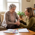 Two women share a comforting moment: one elderly woman holds the other's hand in a sunny living room with a plant and photos on a shelf nearby.” ,