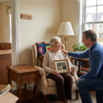 Older woman sitting in a chair holding a framed family photo while a man kneels beside her in a living room with moving boxes nearby.